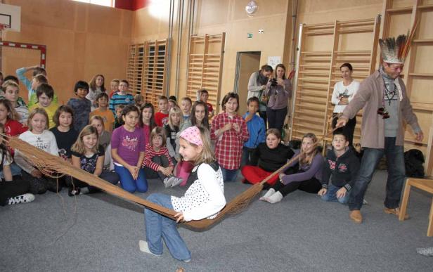 Eine Gruppe von Kindern sitzt auf dem Boden, während ein Mann mit Federschmuck eine Schnur hält.