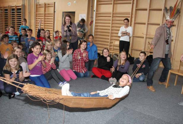 Ein Mann mit Federschmuck zieht ein Mädchen in einer Hängematte vor einer Gruppe von Kindern.