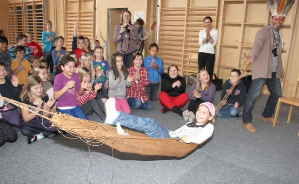 Ein Mädchen liegt in einer Hängematte, während eine Gruppe von Kindern und ein Mann mit Kopfschmuck applaudieren.