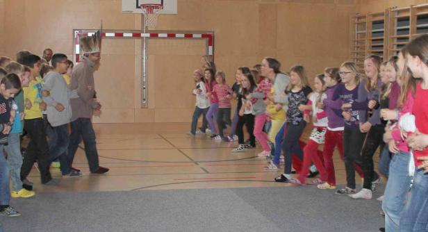 Eine Gruppe Kinder tanzt in einer Turnhalle, angeführt von einem Mann mit Federschmuck.