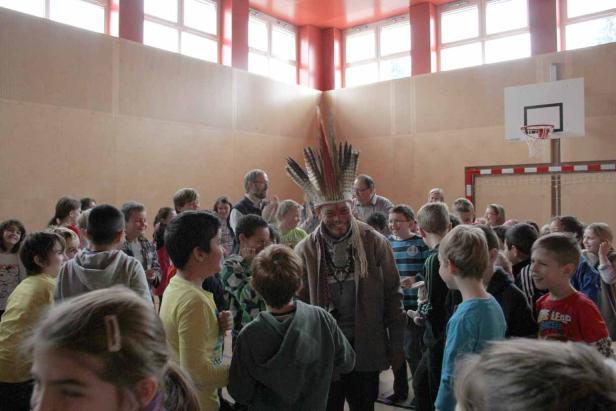 Ein Mann mit Federschmuck steht in einer Turnhalle umringt von Kindern.