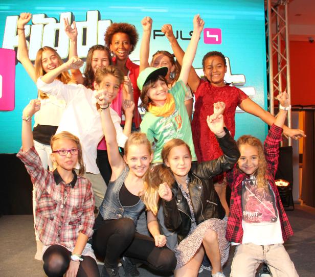 Eine Gruppe von Kindern und eine Frau posieren mit erhobenen Fäusten vor einem Bildschirm mit der Aufschrift „Kids“.