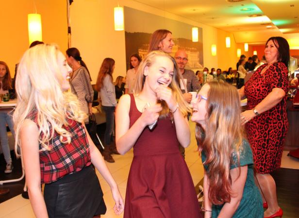 Drei lachende junge Frauen stehen in einem Raum, im Hintergrund weitere Personen.