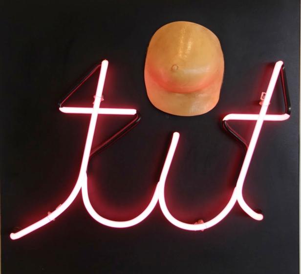 Eine Neonreklame zeigt das Wort „tit“ neben einer Silikonbrust.