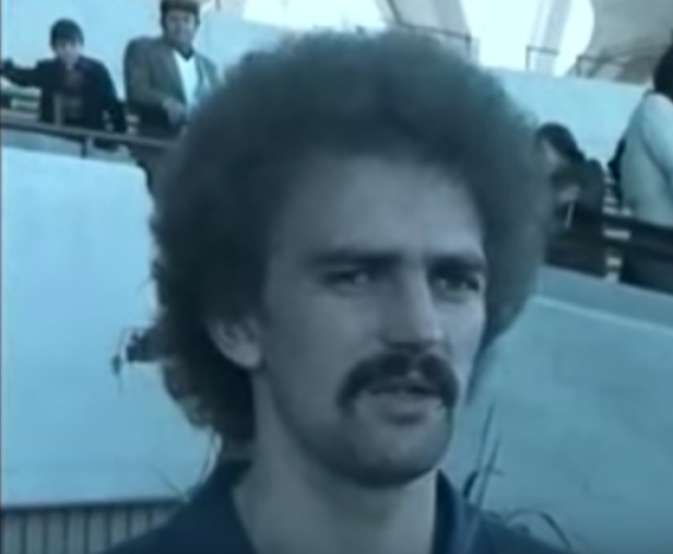 Ein Mann mit Afro und Schnurrbart steht vor einer Tribüne mit Zuschauern.