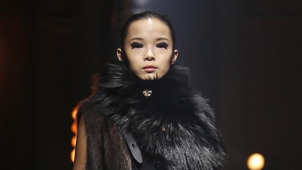 Eine Frau mit dunklem Haar und auffälligem Make-up trägt einen Pelzkragen.