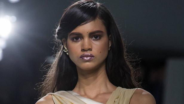 Eine Frau mit dunklen Haaren und glitzerndem Lippenstift trägt ein helles Kleid.