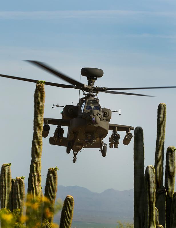 Ein Apache-Kampfhubschrauber fliegt über eine Wüstenlandschaft mit Kakteen.