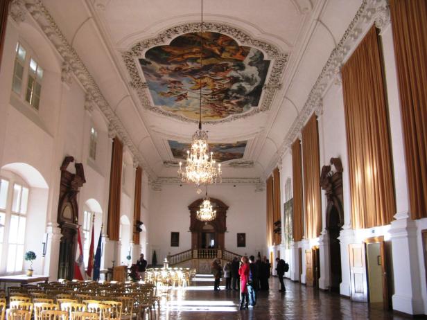 Ein großer Saal mit Deckenmalereien und Kristalllüstern, in dem sich Menschen aufhalten.