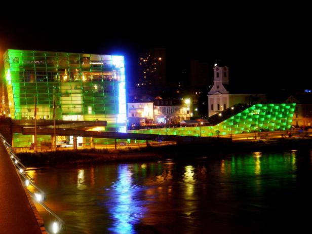 Das Ars Electronica Center und die Stadtpfarrkirche in Linz bei Nacht, beleuchtet in Grün.