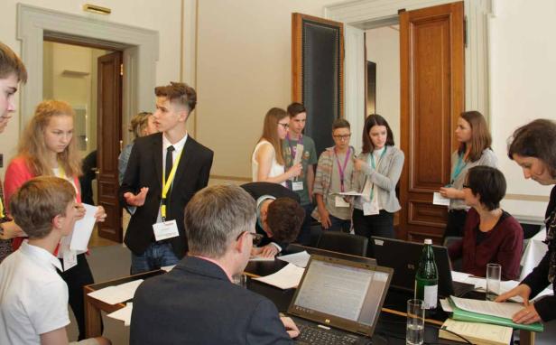 Eine Gruppe junger Leute steht in einem Raum und diskutiert, einige mit Laptops vor sich.