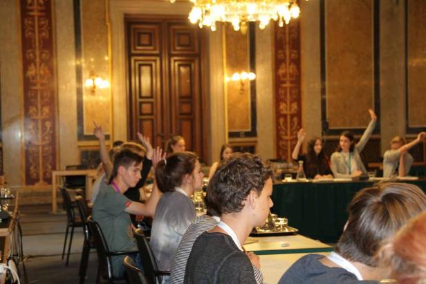 In einem reich verzierten Saal heben mehrere junge Leute ihre Hände.