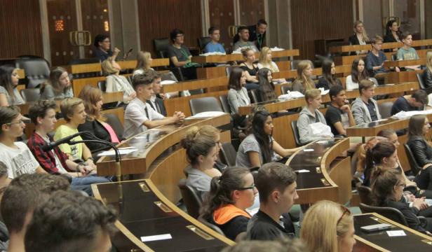 Eine Gruppe junger Menschen sitzt in einem großen Saal mit Holzbänken und Tischen.