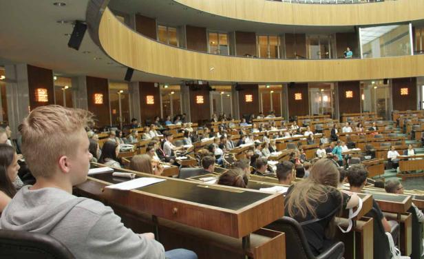 Zahlreiche Menschen sitzen in einem großen Saal mit hölzernen Tischen und Stühlen.