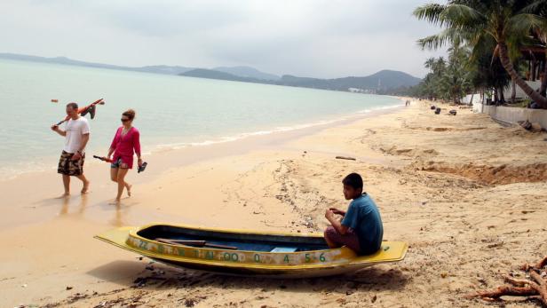 Ein Junge sitzt in einem gelben Boot am Strand, während im Hintergrund ein Paar spaziert.