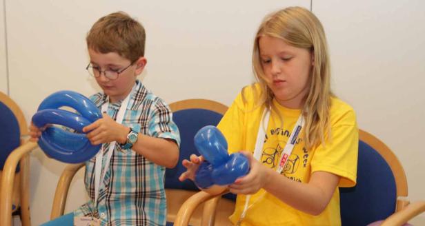Ein Junge und ein Mädchen sitzen auf Stühlen und formen blaue Luftballons.