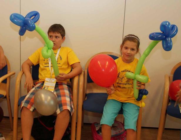 Ein Junge und ein Mädchen sitzen mit Luftballons in Form von Blumen und einem roten Ballon da.