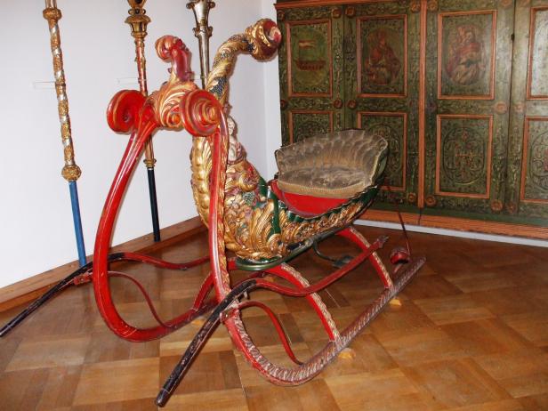 Ein reich verzierter, roter und goldener Schlitten steht auf einem Holzboden.