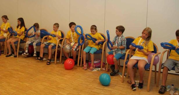 Eine Gruppe Kinder sitzt auf Stühlen und formt blaue Luftballons.