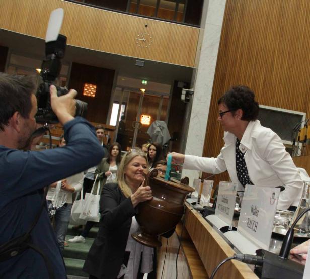 Eine Frau hält eine hölzerne Urne, während eine andere Frau einen Gegenstand hineinlegt.