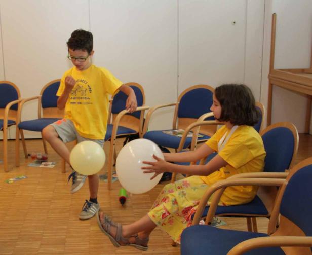 Zwei Kinder spielen mit Ballons in einem Raum voller Stühle.