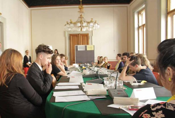 Eine Gruppe junger Leute sitzt an einem langen Tisch mit Mikrofonen in einem Konferenzraum.