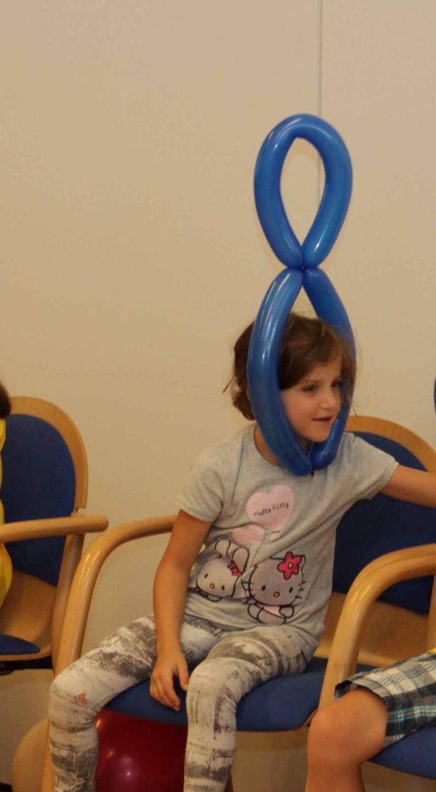 Ein Mädchen sitzt mit einem blauen Ballon um den Kopf auf einem Stuhl.