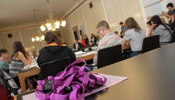 Teilnehmer sitzen an Tischen in einem Konferenzraum, im Vordergrund lilafarbene Schlüsselbänder.
