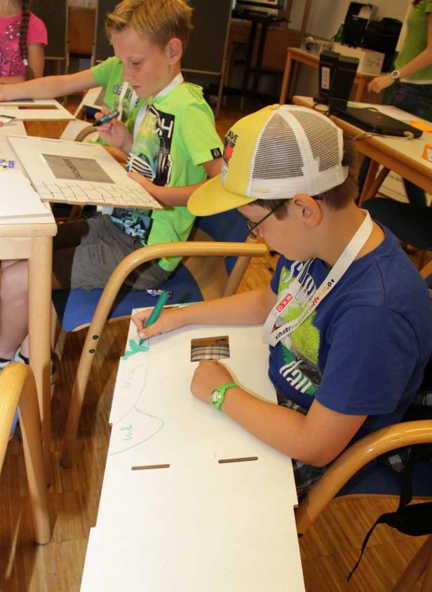 Kinder sitzen an Tischen und malen mit Stiften auf Papier.