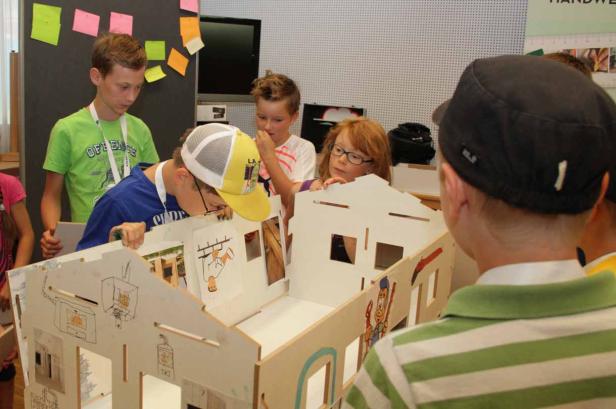 Eine Gruppe Kinder betrachtet ein Modellhaus mit Zeichnungen an den Wänden.