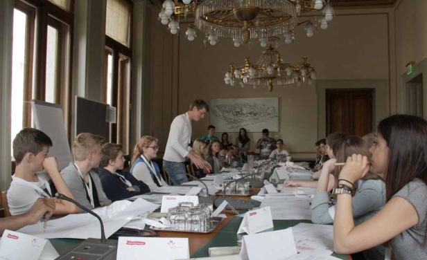 Jugendliche sitzen an einem Tisch in einem großen Raum mit Kronleuchtern.