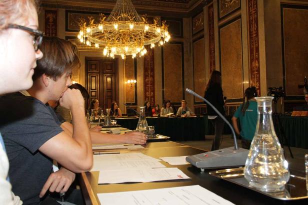 Personen sitzen an einem Tisch in einem opulenten Saal unter einem Kronleuchter.