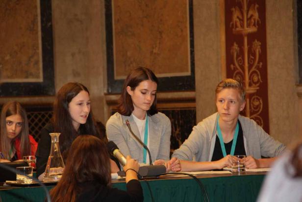 Eine Gruppe junger Leute sitzt an einem Tisch mit Mikrofonen, möglicherweise bei einer Konferenz oder Podiumsdiskussion.