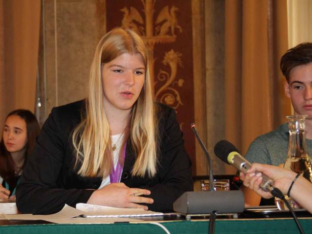 Eine junge Frau mit blonden Haaren spricht bei einer Konferenz am Mikrofon.