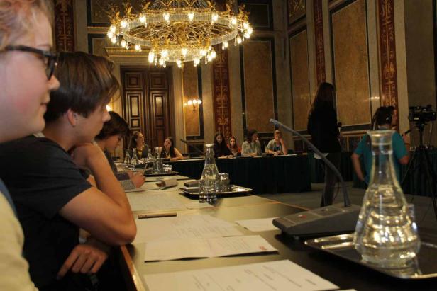 Personen sitzen an Tischen in einem opulenten Saal unter einem Kronleuchter.