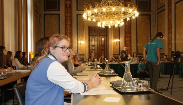Eine junge Frau mit Brille sitzt an einem Tisch während einer Konferenz in einem prunkvollen Saal.