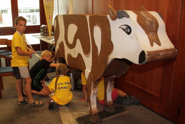 Kinder melken eine bemalte Kuh aus Pappe.