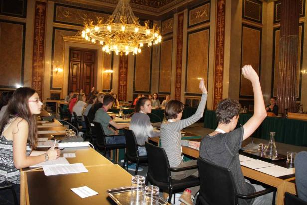 In einem prunkvollen Saal heben mehrere Personen die Hände.