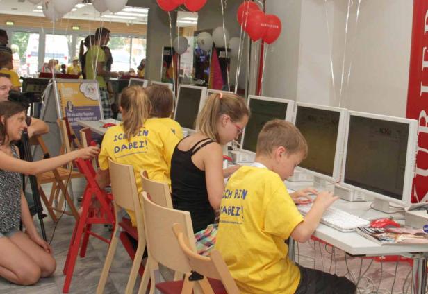 Kinder sitzen an Computern und tragen gelbe T-Shirts mit der Aufschrift „Ich bin dabei!“.