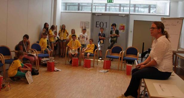 Eine Gruppe Kinder sitzt und steht in einem Raum, während eine Frau spricht.