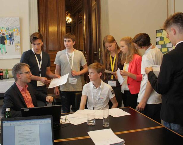 Eine Gruppe Jugendlicher steht um einen Tisch, an dem ein Mann mit Laptop sitzt.