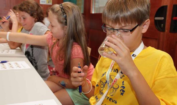 Ein Junge mit Brille trinkt Wasser, während andere Kinder an Tischen sitzen und arbeiten.