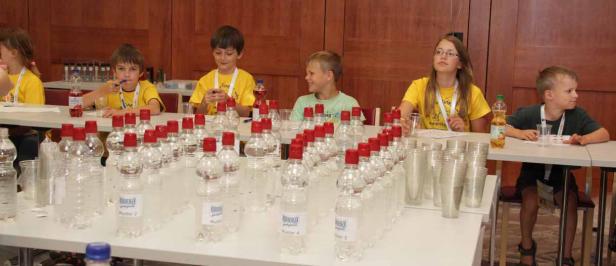Kinder sitzen an Tischen mit Wasserflaschen und Plastikbechern bei einer Veranstaltung.