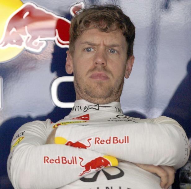 Sebastian Vettel mit verschränkten Armen und Stirnrunzeln vor einem Red Bull Hintergrund.