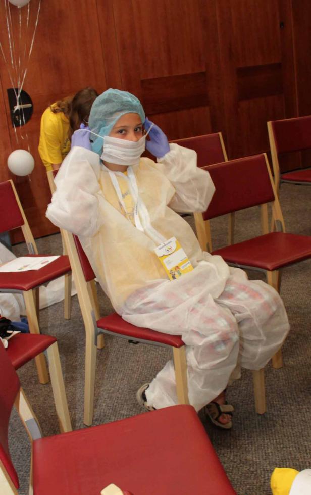 Ein Kind sitzt mit Haube, Maske, Handschuhen und Schutzkittel auf einem Stuhl.