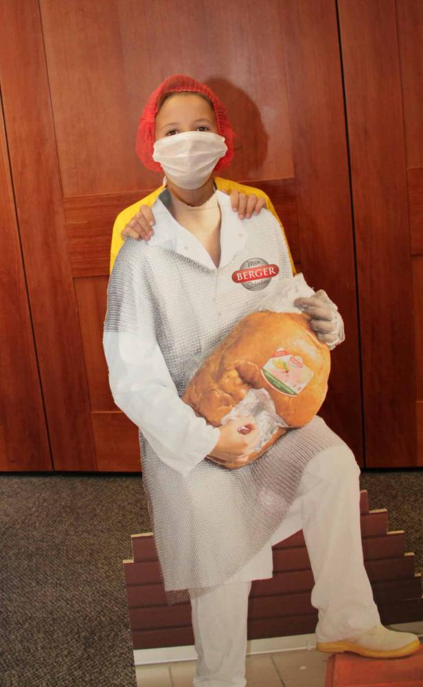 Ein Kind mit rotem Haarnetz und Mundschutz hält eine Pappfigur eines Metzgers mit einem Schinken in den Armen.