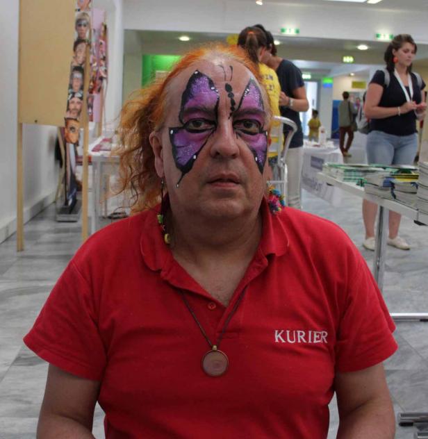 Ein Mann mit rotem Haar und Schmetterlingsgesichtsbemalung trägt ein rotes „Kurier“-Polohemd.