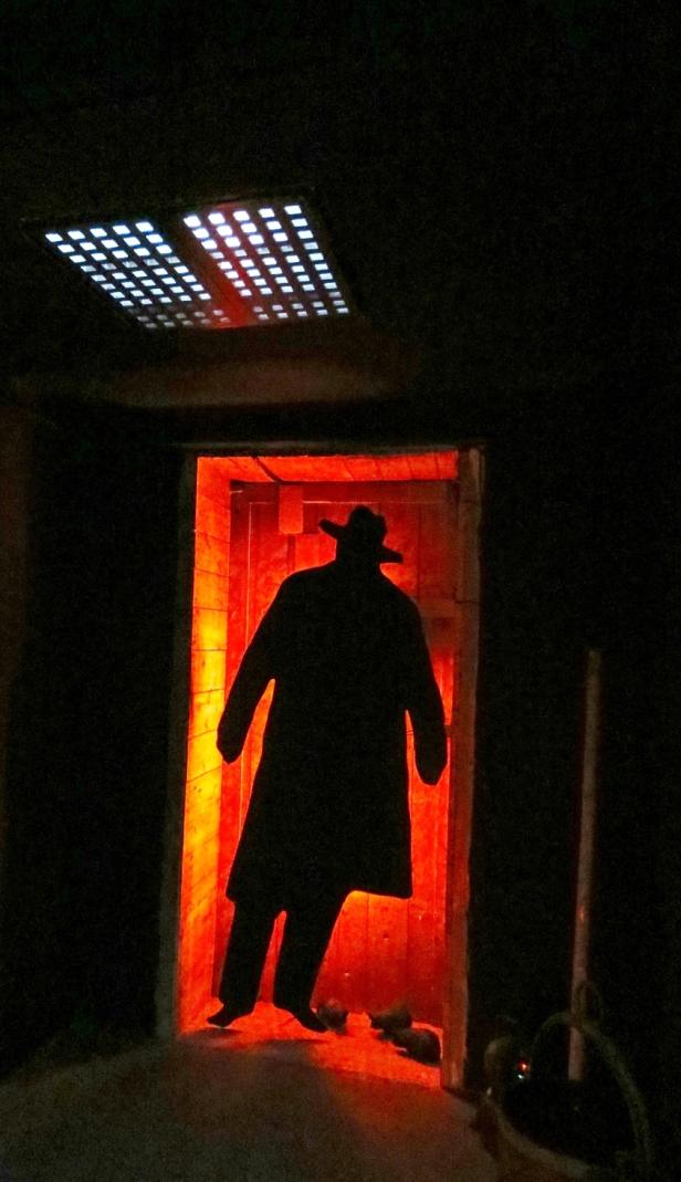 Die Silhouette eines Mannes mit Hut und Mantel steht in einer rot beleuchteten Türöffnung.