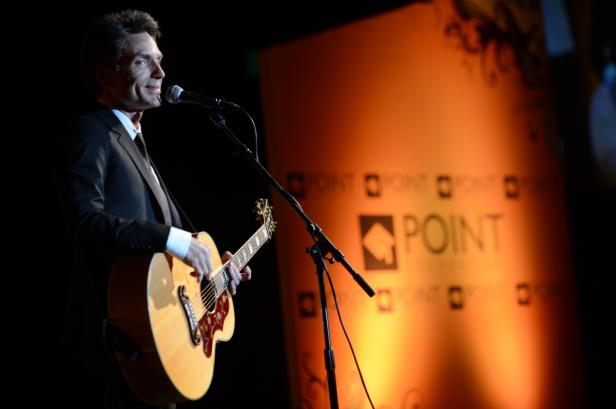 Ein Mann im Anzug spielt Gitarre und singt in ein Mikrofon vor einem orangefarbenen Hintergrund.