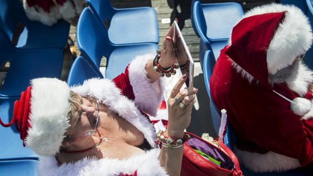Zwei als Weihnachtsmann verkleidete Personen sitzen auf blauen Stühlen, eine davon benutzt ein Tablet.